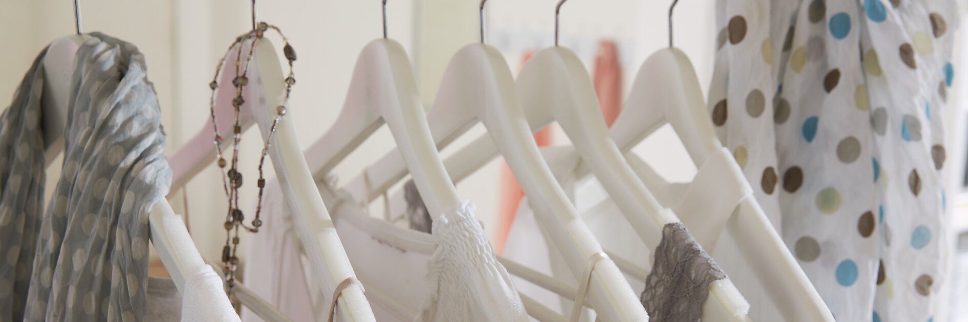 A row of white clothes on white hangers is displayed on a metal rod. A necklace with beads hangs on one hanger, and a polka dot scarf drapes over the rod. The background is softly blurred.