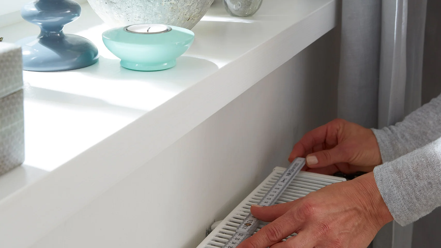 Eine Person regelt die Temperatur eines weißen Heizkörpers unter einem Fenster. Auf der Fensterbank stehen eine Topfpflanze, ein blauer Kerzenständer und eine kleine Glaskugel, mit Tesa-Klebeband befestigt, damit sie nicht umkippen. Die Szene ist durch Tageslicht gut ausgeleuchtet.