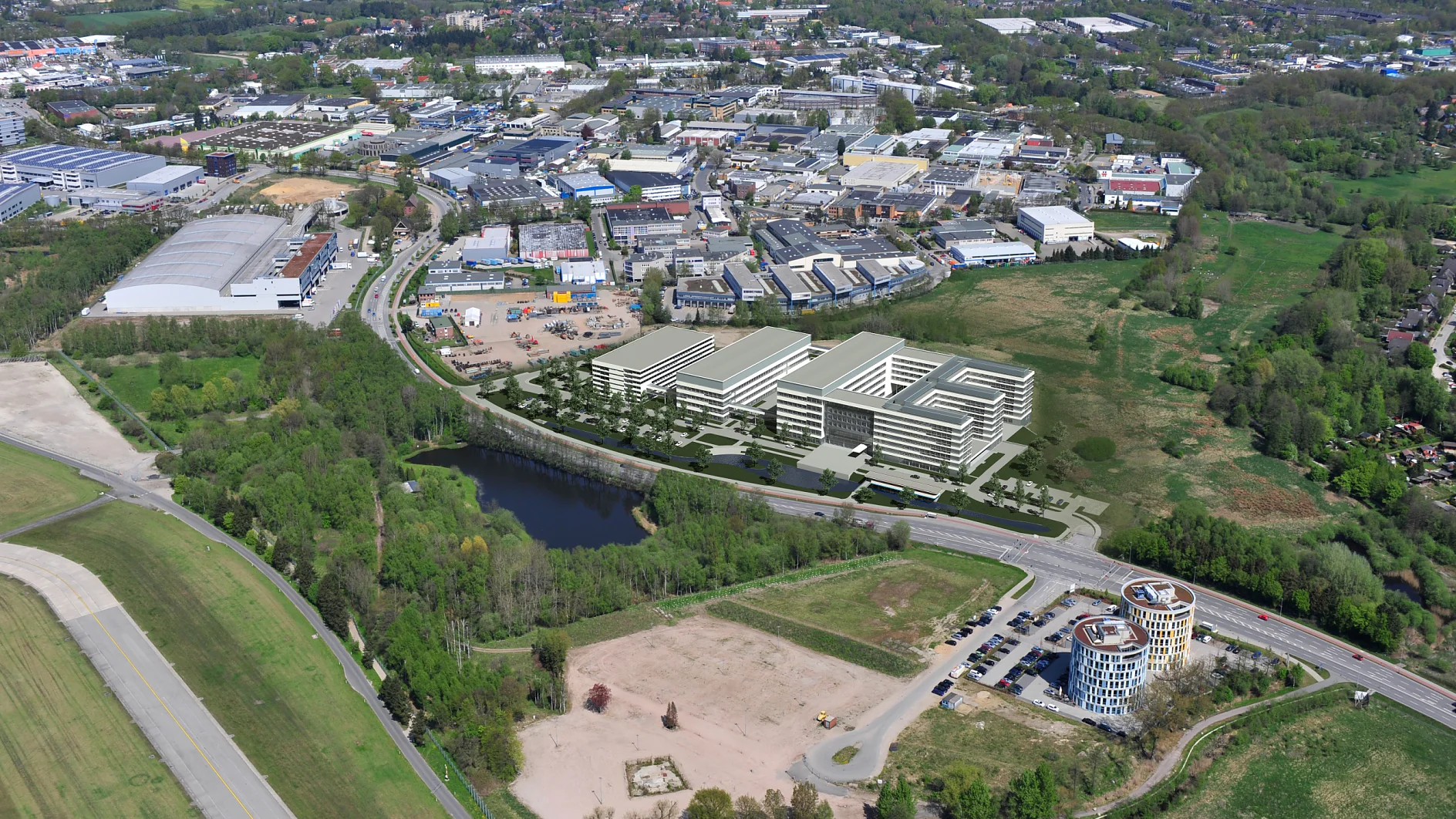 Luftaufnahme eines Industriegebiets mit mehreren Gebäuden, umgeben von Grün. Eine kurvenreiche Straße führt an einem kleinen See und offenen Feldern vorbei. Im Vordergrund steht ein zylindrisches Gebäude in der Nähe eines Parkplatzes. In der Ferne sind Wohngebiete zu sehen.