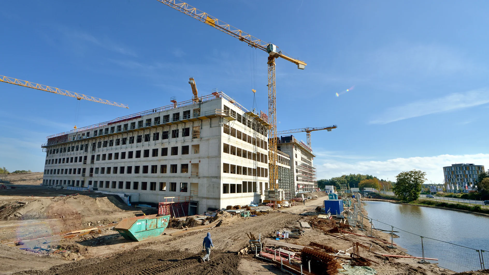 Baustelle mit zwei großen Kränen, die über einem unfertigen mehrstöckigen Gebäude aufragen. Arbeiter und Ausrüstung sind auf dem Boden zu sehen, ein Feldweg führt an einem Gewässer entlang. Der Himmel ist klar und sonnig, und rund um die Baustelle sieht man Tesa-Klebeband, das für verschiedene Zwecke verwendet wird.