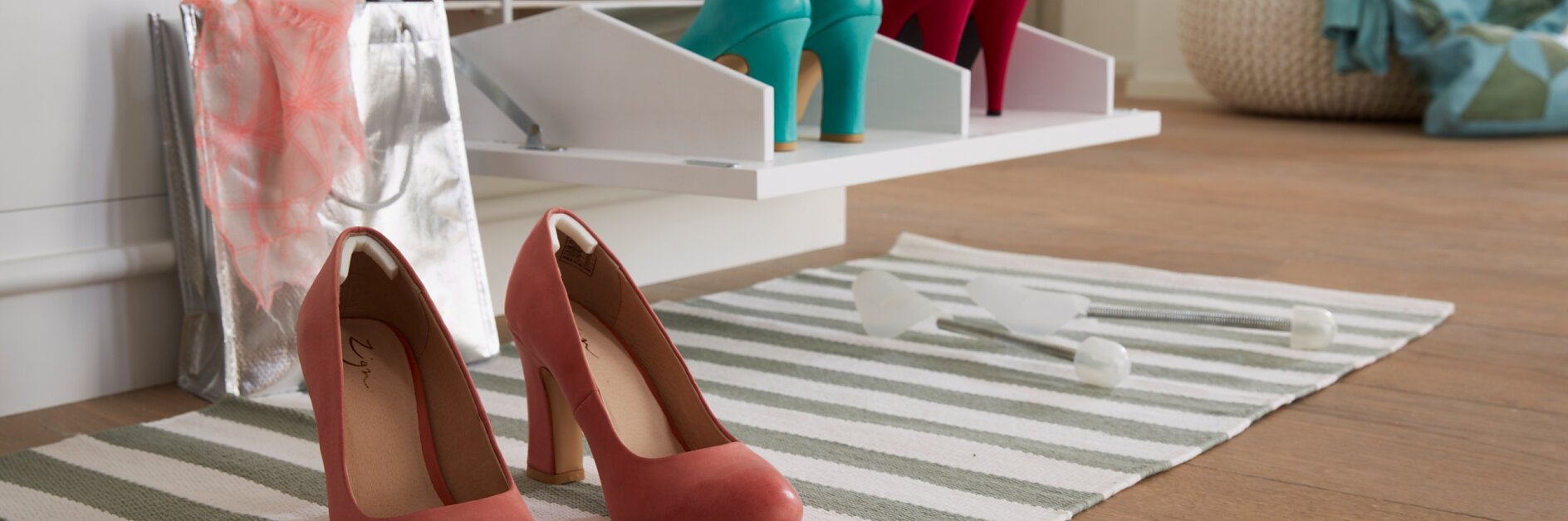 Red high-heeled shoes on a striped rug with a shoe organizer in the background holding teal and red shoes. Two tesa tape shoehorns and shopping bags are on the floor nearby. A pouf and pillows are in the corner of the room.