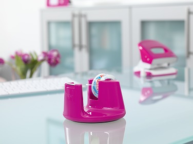 Auf einem Glasschreibtisch steht ein rosafarbener Tesa-Klebebandspender. Im Hintergrund sind ein unscharfer rosafarbener Locher und lilafarbene Blumen auf einem Schrank zu sehen. Die Szene vermittelt ein helles und farbenfrohes Büroambiente.