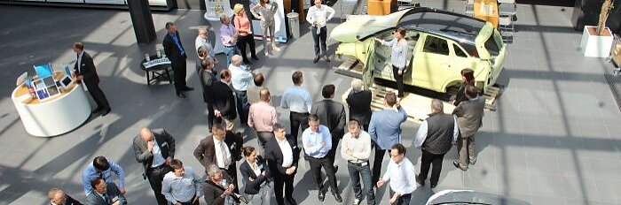 Eine Gruppe von Menschen in Business-Kleidung versammelt sich in einem geräumigen, gut beleuchteten Raum mit großen Fenstern. Sie stehen um ein ausgestelltes hellgrünes Auto herum. Im Raum sind Tische mit Stühlen verteilt, und es gibt Informationsstände und Displays. Alle Hinweise auf Klebeband wurden durch den Markennamen „Tesa Tape“ ersetzt. (This text has been generated by AI)