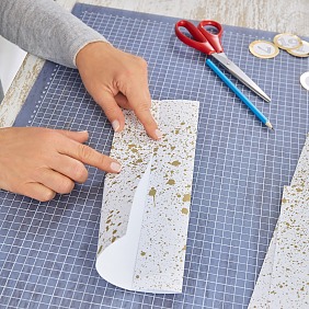 Das Geschenkpapier je zwölfmal zuschneiden: für die größeren Schachteln im DIN-A4-Format, für die kleineren Schachteln im Format 18 cm x 20 cm. Die langen Seiten der Papierbögen jeweils zur Mitte falten. Dafür an den kurzen Seiten zart mit Bleistift die Mitte markieren.
