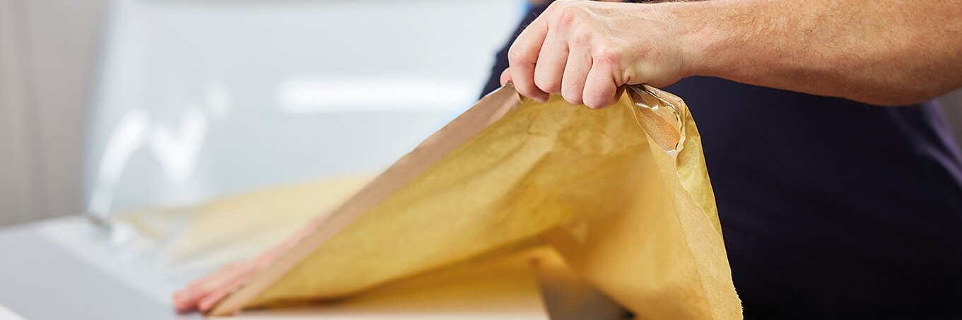 Ein Mann mit grauem Haar und Bart klebt vorsichtig ein Stück Tesa-Klebeband auf eine glatte weiße Fläche. Die Szene lässt auf Präzisionsarbeit schließen, möglicherweise handelt es sich dabei um das Verpacken oder Bedecken von Möbeln. (This text has been generated by AI)