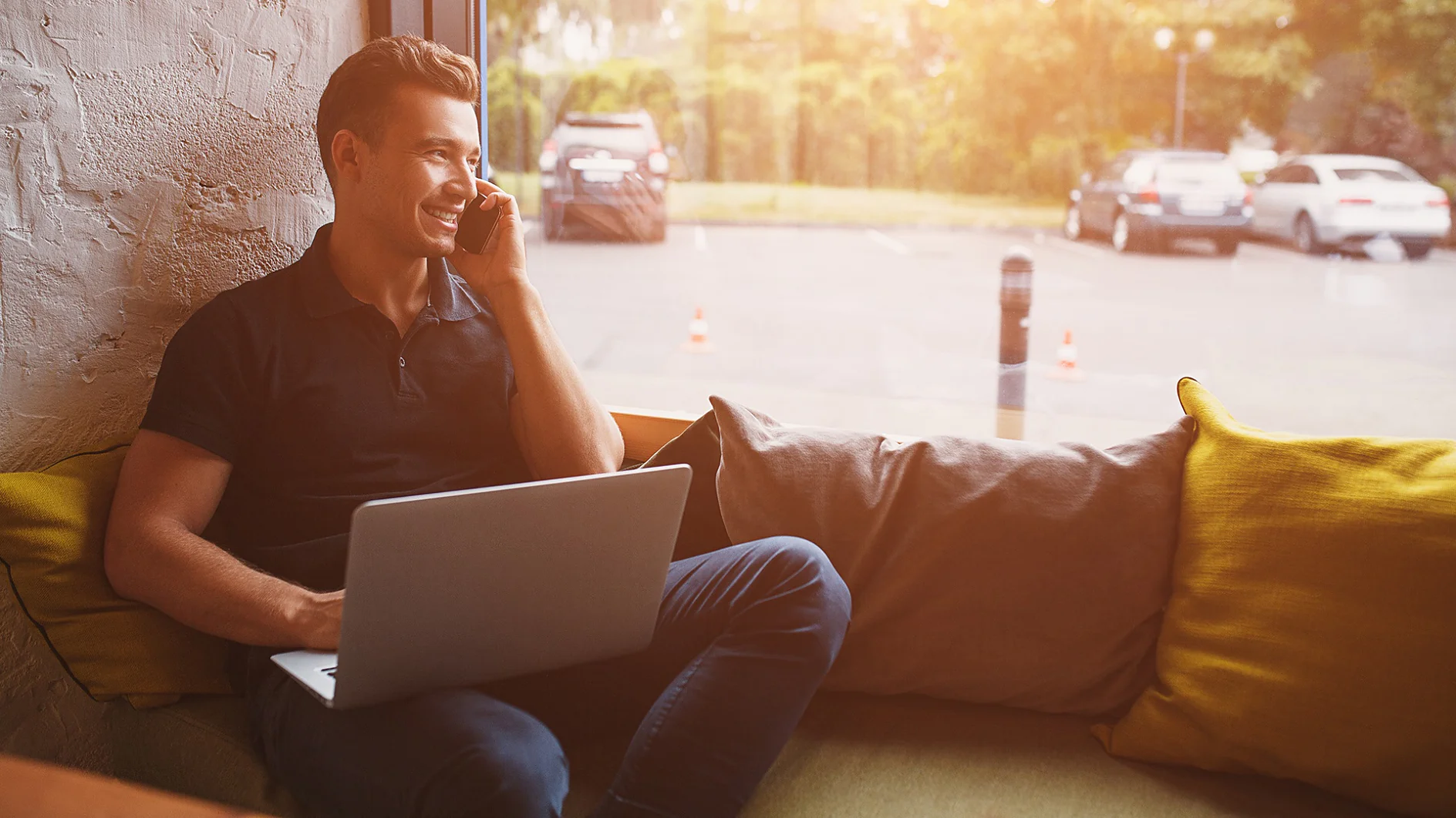 Ein Mann sitzt auf einer gepolsterten Bank neben einem Fenster, hält ein Telefon ans Ohr und lächelt. Auf seinem Schoß liegt ein geöffneter Laptop. Sonnenlicht fällt durch das Fenster und erhellt den Raum. Draußen sind geparkte Autos zu sehen. (This text has been generated by AI)