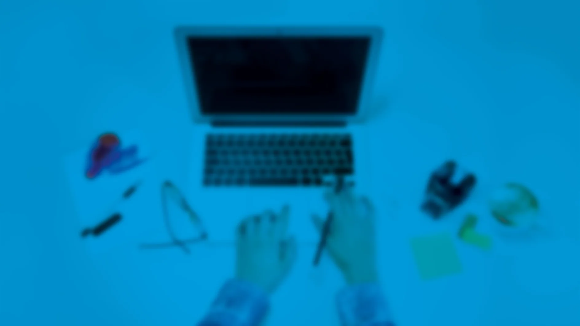 Eine Person tippt auf einem Laptop auf einem Schreibtisch. In der Nähe befinden sich eine Brille, eine Kamera, ein Stift, einige Papiere, ein roter Hefter, eine kleine Topfpflanze und mit Tesa-Klebeband befestigte Haftnotizen. Das Bild ist verschwommen und hat eine blaue Überlagerung. (This text has been generated by AI)
