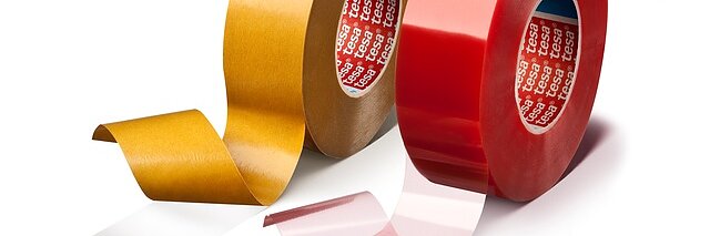 Ausgestellt sind zwei Rollen Tesa-Klebeband. Die linke Rolle ist goldfarben mit einem teilweise abgerollten transparenten Streifen. Die rechte Rolle ist rot mit einem ebenfalls abgerollten transparenten Streifen. Auf beiden Rollen sind in der Mitte die Markenkerne sichtbar. (This text has been generated by AI)
