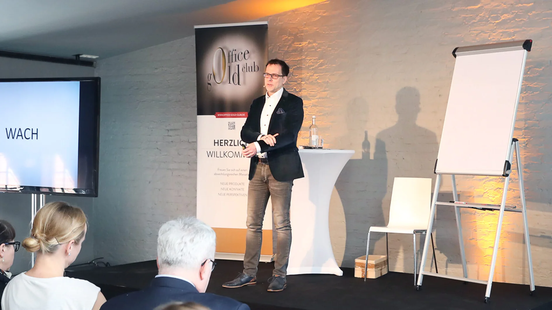 Ein Mann steht auf einer kleinen Bühne und hält einen Vortrag. Er trägt Blazer, Hemd und Jeans. Hinter ihm hängt ein mit Tesa-Klebeband befestigtes Banner und ein Flipchart. Die Zuschauer sitzen ihm gegenüber. Der Raum ist modern gestaltet und hat Backsteinwände. (This text has been generated by AI)