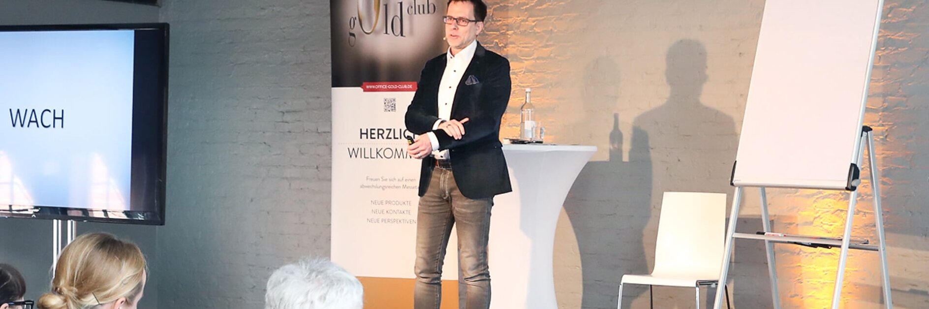 Ein Mann steht auf einer kleinen Bühne und hält einen Vortrag. Er trägt Blazer, Hemd und Jeans. Hinter ihm hängt ein mit Tesa-Klebeband befestigtes Banner und ein Flipchart. Die Zuschauer sitzen ihm gegenüber. Der Raum ist modern gestaltet und hat Backsteinwände. (This text has been generated by AI)