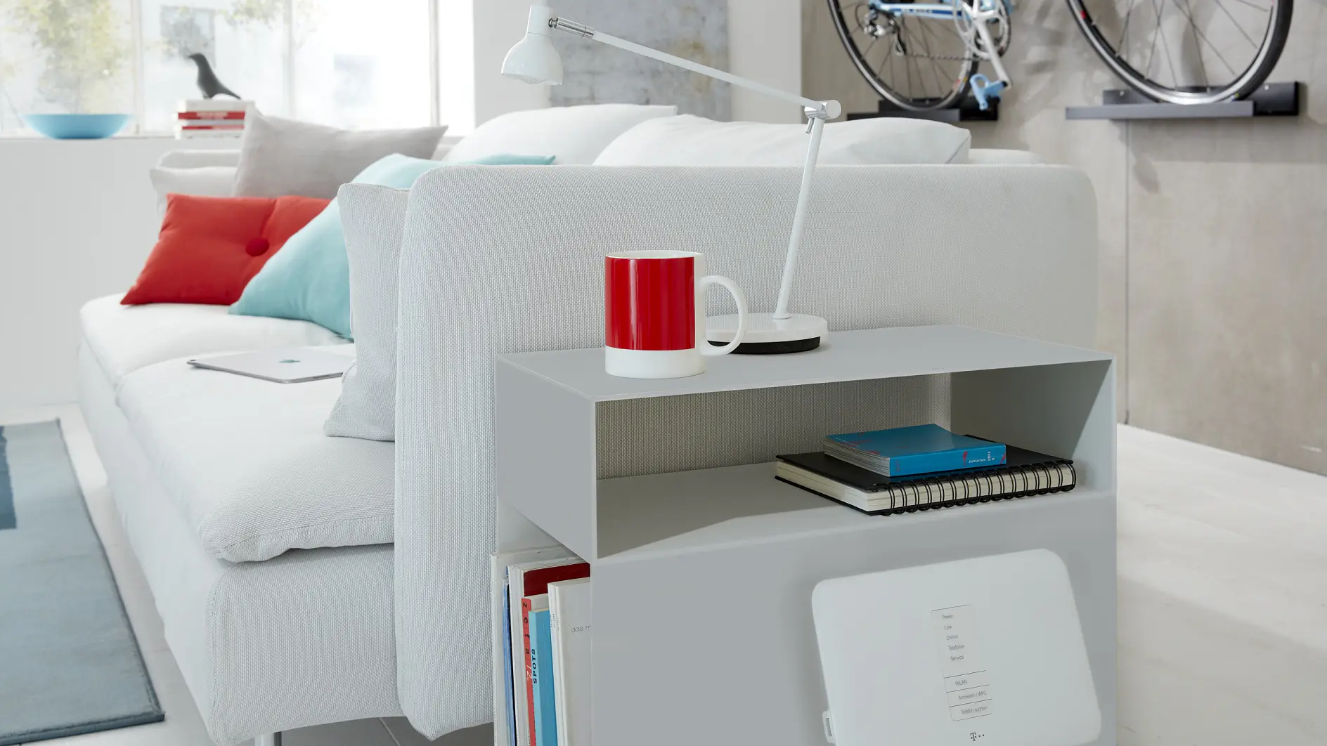 A modern living room showcases a white sofa adorned with red and gray throw pillows. A red mug and a white lamp are placed on a side table. A bicycle is mounted on the wall using tesa tape. Books and a notepad are neatly stored on the table shelf.