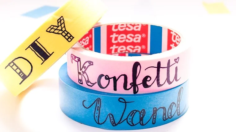 Drei Rollen buntes Tesa-Klebeband übereinander gestapelt. Die obere Rolle ist gelb und trägt die Aufschrift „DIY“, die mittlere Rolle ist rosa und trägt die Aufschrift „Konfetti“ und die untere Rolle ist blau und trägt die Aufschrift „Wand“. Im Inneren der rosa Rolle ist ein Markenlabel von Tesa zu sehen. (This text has been generated by AI)