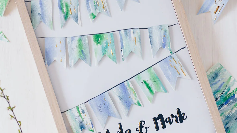 Ein Holzrahmen zeigt einen weißen Hintergrund mit blauen und grünen Aquarellpapierfahnen, die zwei Girlanden bilden. Der Text „Nikola & Mark“ ist in schwarzer Schreibschrift unten geschrieben. Dekorative Zweige mit Blumen sind um den Rahmen gelegt und sorgfältig mit Tesaband befestigt. (This text has been generated by AI)