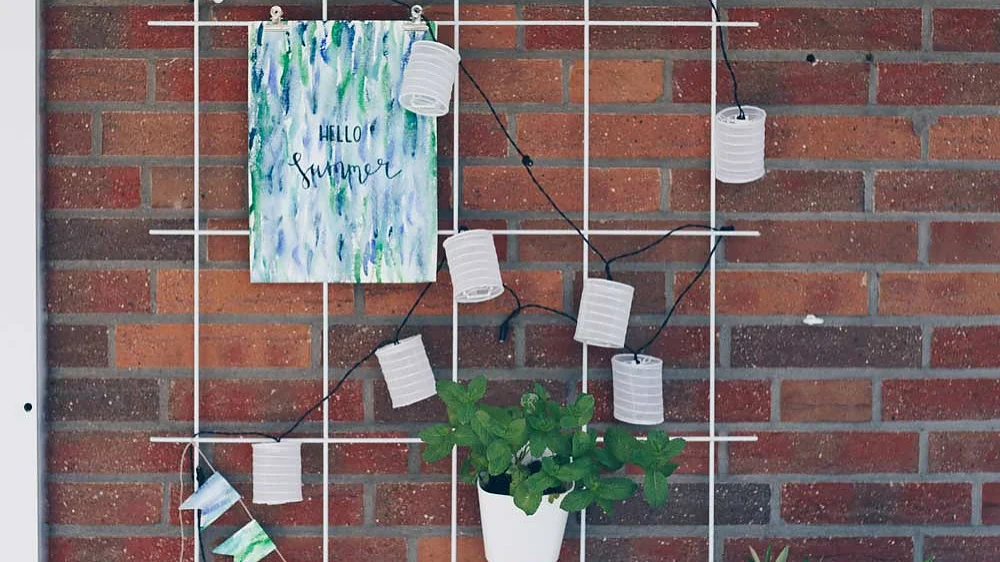 An einer Backsteinmauer hängt ein weißes Metallgitter mit Lichterketten, weißen Laternen, einer Topfpflanze und einem kleinen Banner. Über der Pflanze hängt ein Schild mit der Aufschrift „Hallo Sommer“. Zwei weitere Topfpflanzen sind unten angebracht und mit Tesa-Klebeband sicher befestigt. (This text has been generated by AI)