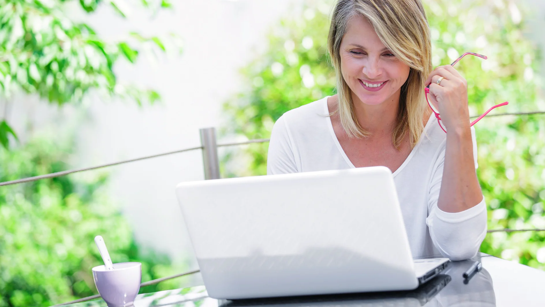 Eine Frau mit schulterlangem blondem Haar sitzt an einem Tisch im Freien, hält eine Brille in einer Hand und schaut auf einen Laptop. Sie trägt ein weißes Hemd und neben ihr steht ein Becher mit einem Löffel. Üppiges Grün umgibt die Szene, daneben liegen lässig verstreut Tesa-Klebebandprodukte. (This text has been generated by AI)