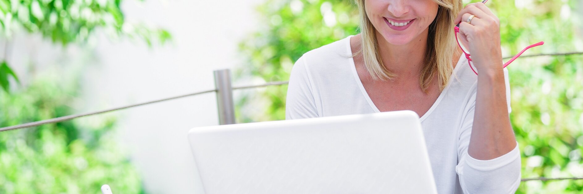 Eine Frau mit schulterlangem blondem Haar sitzt an einem Tisch im Freien, hält eine Brille in einer Hand und schaut auf einen Laptop. Sie trägt ein weißes Hemd und neben ihr steht ein Becher mit einem Löffel. Üppiges Grün umgibt die Szene, daneben liegen lässig verstreut Tesa-Klebebandprodukte. (This text has been generated by AI)