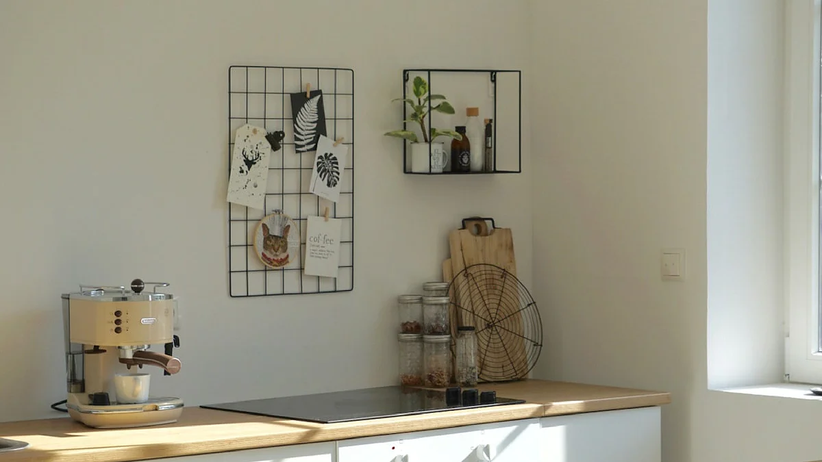 Eine minimalistische Küche mit heller Holzarbeitsplatte und weißen Schränken. Auf der Arbeitsplatte stehen eine Kaffeemaschine, Gläser und Schneidebretter. Ein Drahtgitter mit mit Tesa-Klebeband befestigten Fotos und ein kleines Regal mit Flaschen schmücken die weiße Wand. Natürliches Licht fällt von rechts ein. (This text has been generated by AI)