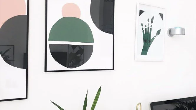 An einer minimalistischen weißen Wand hängen drei gerahmte Kunstwerke mit abstrakten geometrischen Formen und Pflanzenmotiven, die mit Tesa-Klebeband sicher aufgehängt sind. Darunter sind eine grüne Pflanze und ein Computermonitor auf einem Schreibtisch neben einer Schreibtischlampe aus Messing zu sehen. (This text has been generated by AI)