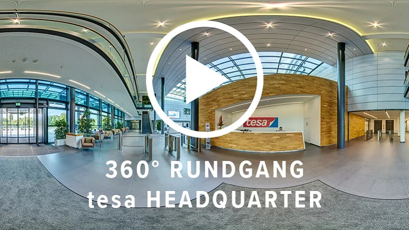 Ein Panoramablick auf die Lobby des tesa-Hauptsitzes. Das Bild zeigt ein modernes Interieur mit einem Glaseingang, einem Empfangstresen mit dem tesa-Klebeband-Logo und Sitzbereichen. Der Text auf dem Bild lautet „360° RUNDGANG tesa HEADQUARTER“ mit einem Play-Button-Symbol. (This text has been generated by AI)