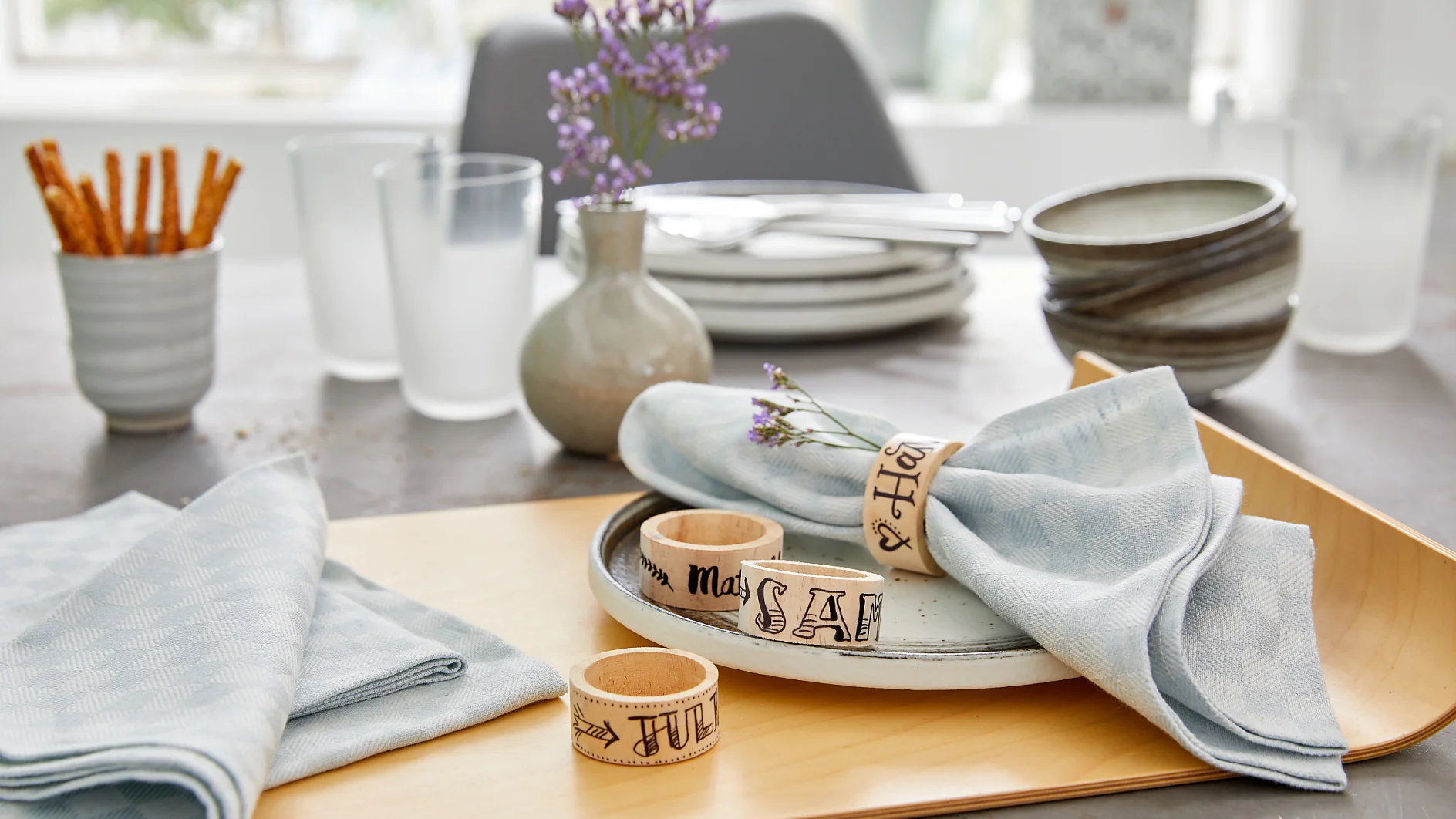 A table is set with light blue napkins, each wrapped in a wooden ring with a month name. Plates are stacked, and bowls are adjacent. Twigs of lavender are placed on two plates. Glasses and a vase are in the background. Pretzel sticks are in a cup. (This text has been generated by AI)