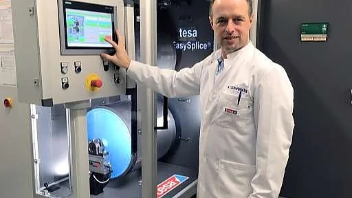 employee splicing machine Eine Person im weißen Laborkittel steht neben einer Maschine mit einem Bildschirm. Auf dem Bildschirm sind mehrere grüne Knöpfe zu sehen. Die Maschine trägt die Aufschrift „tesa EasySplice“. Die Person lächelt und hat eine Hand auf der Maschine. Im Hintergrund ist eine Tür zu sehen. (This text has been generated by AI)