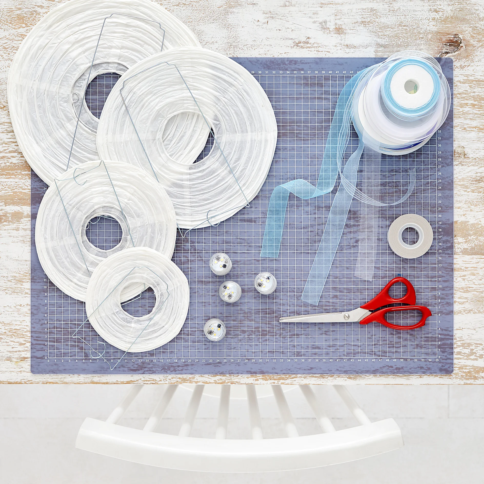 Paper lantern materials are arranged on a cutting mat: circular frames, blue and white ribbons, four tealight candles, scissors, tesa tape, and a spool of ribbon. A white chair is partially visible at the bottom. (This text has been generated by AI)