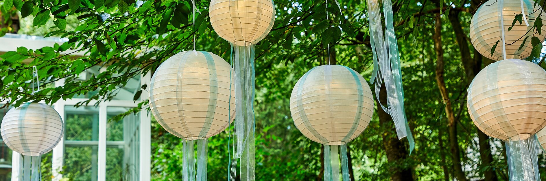Paper lanterns hang from tree branches in an outdoor setting. The lanterns are round and adorned with long, translucent ribbons. Each ribbon is neatly secured using tesa tape to ensure they hang gracefully in the breeze. In the background, there is foliage and a glimpse of building windows. (This text has been generated by AI)