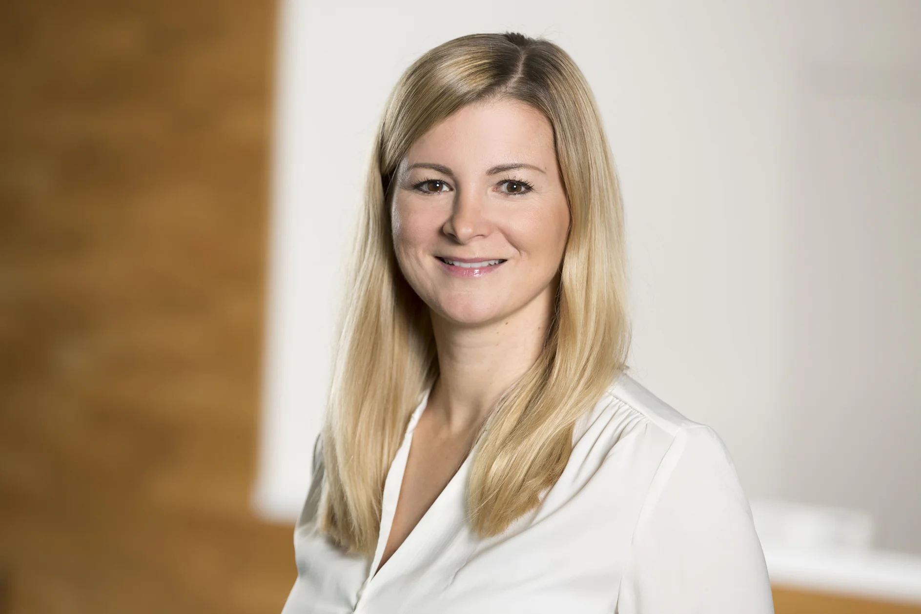 Eine Person mit langen blonden Haaren lächelt und trägt ein weißes Hemd. Der Hintergrund ist mit Braun- und Weißtönen verschwommen. (This text has been generated by AI)