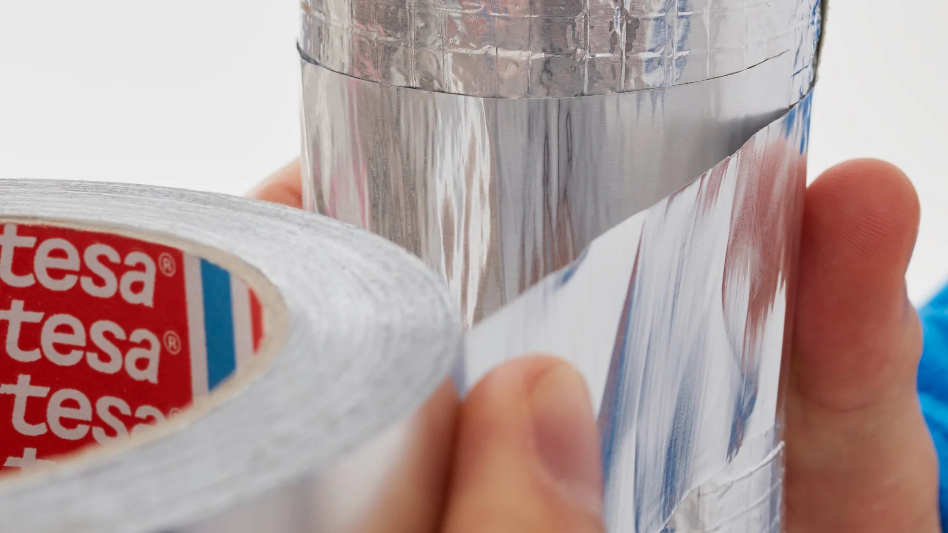 Hände halten einen Streifen reflektierendes, metallisches Tesa-Klebeband von einer Rolle und bringen es an. Die Tesa-Klebebandrolle ist innen mit einem rot-weißen Logo versehen. Die Oberfläche, auf die es geklebt wird, erscheint glänzend und metallisch. (This text has been generated by AI)