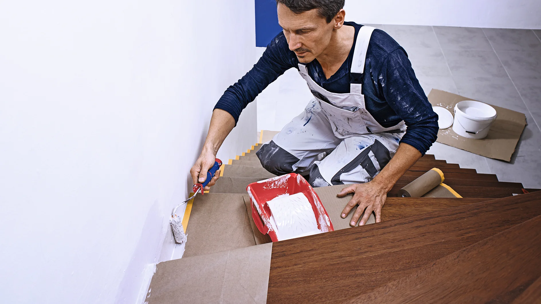 Ein Mann im weißen Overall streicht eine Treppenhauswand mit weißer Farbe. Er kniet auf einer Holztreppe und hält einen Pinsel und eine rote Farbwanne in der Hand. Die Stufen sind zum Schutz mit Pappe abgedeckt und mit Tesa-Klebeband befestigt. Auf einer Stufe daneben stehen Farbdosen.