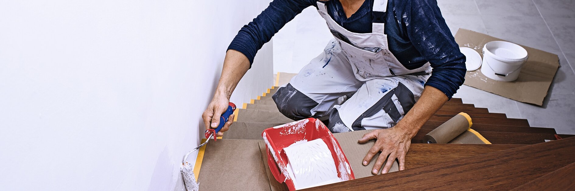 Ein Mann im weißen Overall streicht eine Treppenhauswand mit weißer Farbe. Er kniet auf einer Holztreppe und hält einen Pinsel und eine rote Farbwanne in der Hand. Die Stufen sind zum Schutz mit Pappe abgedeckt und mit Tesa-Klebeband befestigt. Auf einer Stufe daneben stehen Farbdosen. (This text has been generated by AI)