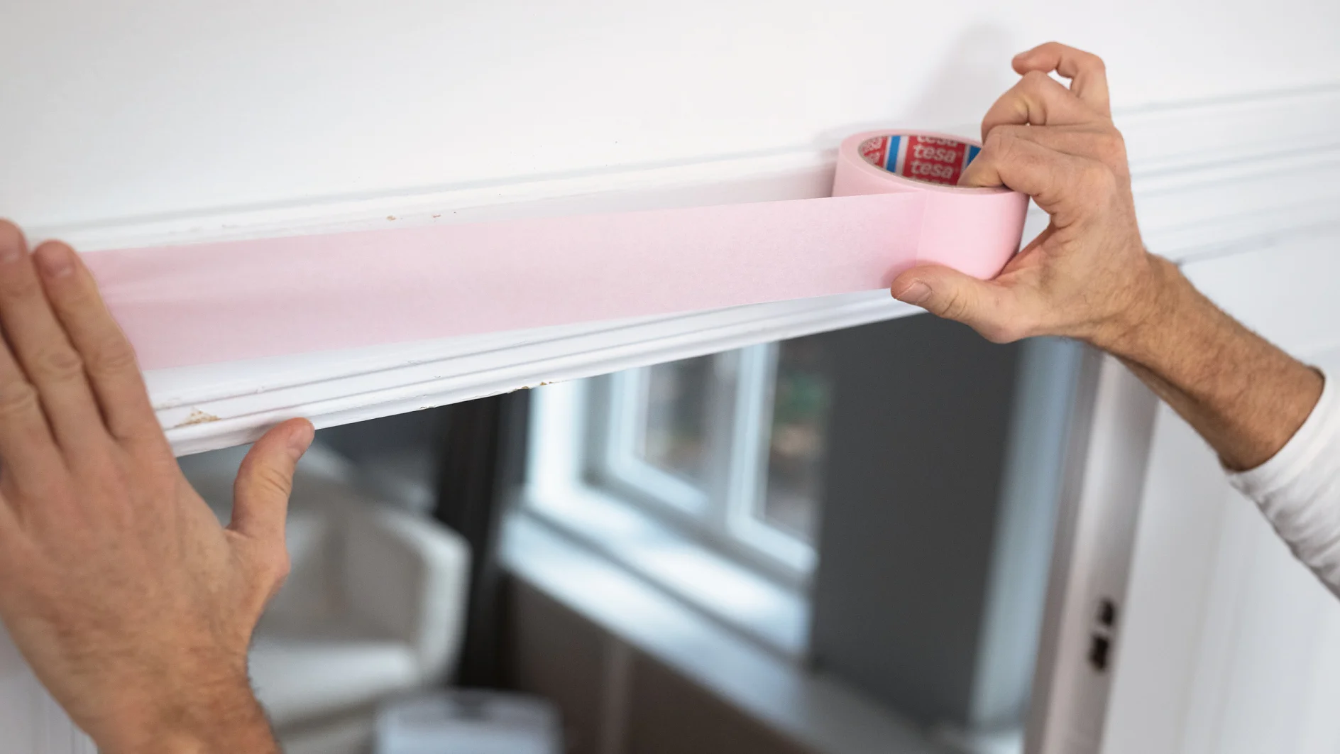 Eine Person klebt rosa Tesa-Klebeband an die Oberkante eines weißen Türrahmens. Durch ein Fenster im Hintergrund fällt Licht ein und beleuchtet das Innere eines Raums mit weißen Wänden und Möbeln.