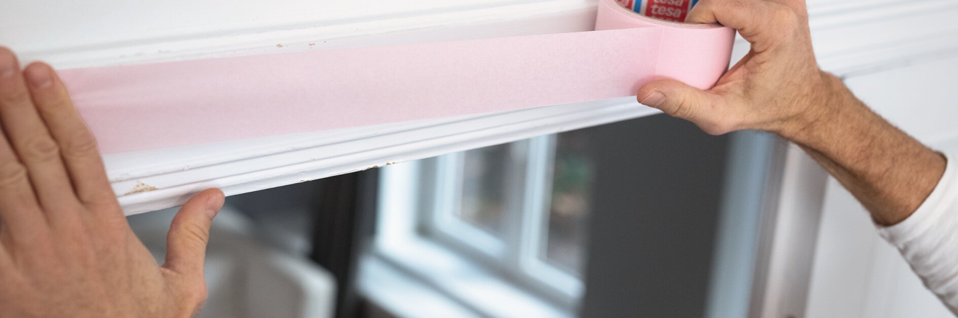 Eine Person klebt rosa Tesa-Klebeband an die Oberkante eines weißen Türrahmens. Durch ein Fenster im Hintergrund fällt Licht ein und beleuchtet das Innere eines Raums mit weißen Wänden und Möbeln. (This text has been generated by AI)
