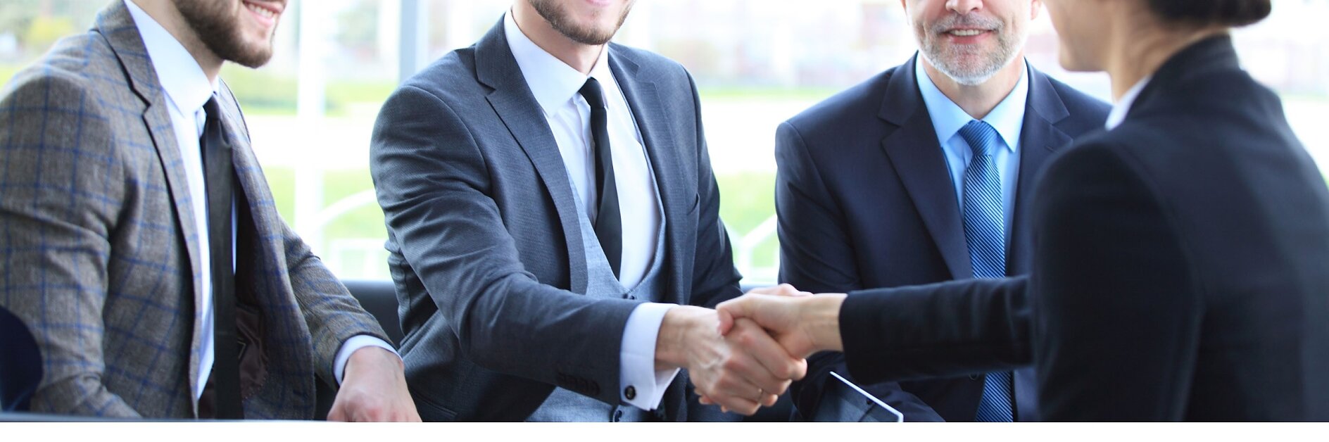 Eine Gruppe von vier Personen in Business-Kleidung sitzt um einen Tisch. Zwei Männer, einer links, der andere rechts, schütteln sich die Hände. Im Hintergrund sind große Fenster zu sehen, durch die man unscharf Tesa-Klebeband im Freien sieht. (This text has been generated by AI)