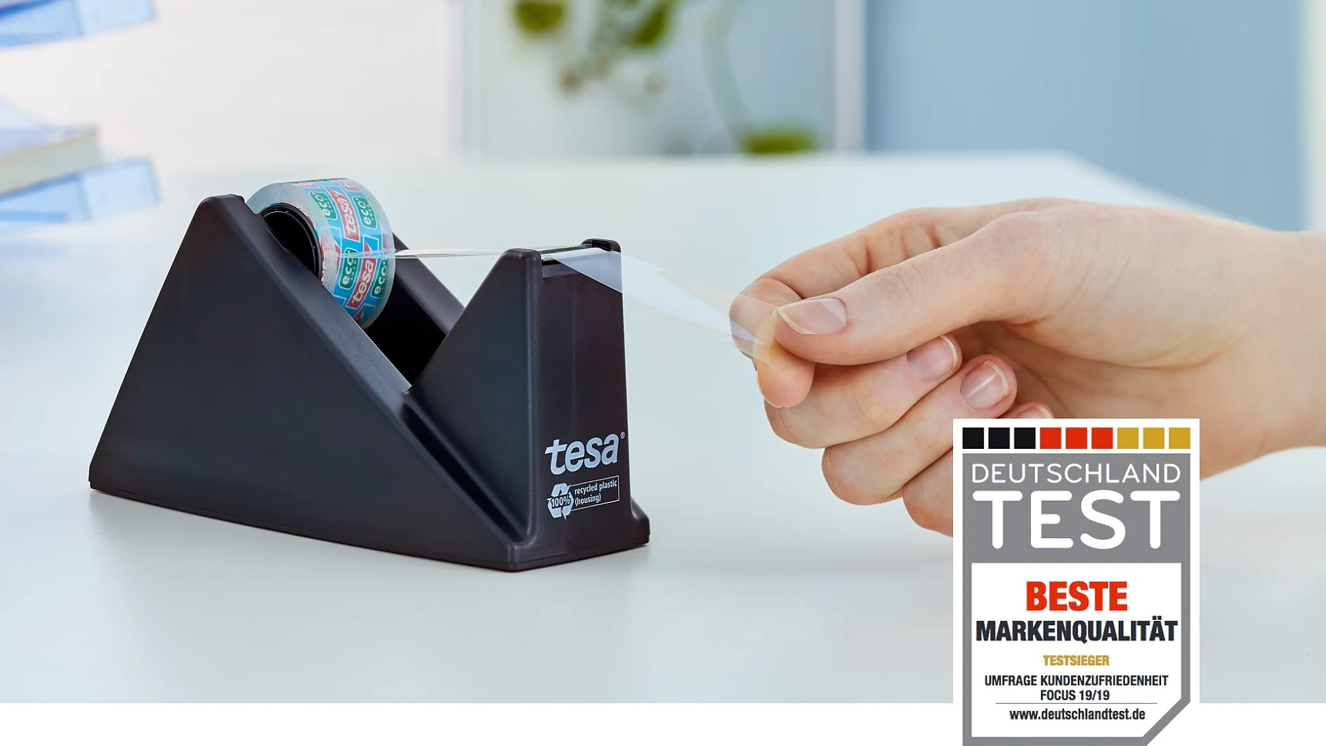 Eine Hand zieht einen Streifen Tesa-Klebeband aus einem schwarzen Klebebandspender mit der Aufschrift „Tesa“. Im Vordergrund ist eine Grafik des Deutschland Test-Preises mit der Auszeichnung „Beste Markenqualität“ zu sehen. Ein unscharfer weißer Hintergrund lässt eine Inneneinrichtung vermuten. (This text has been generated by AI)