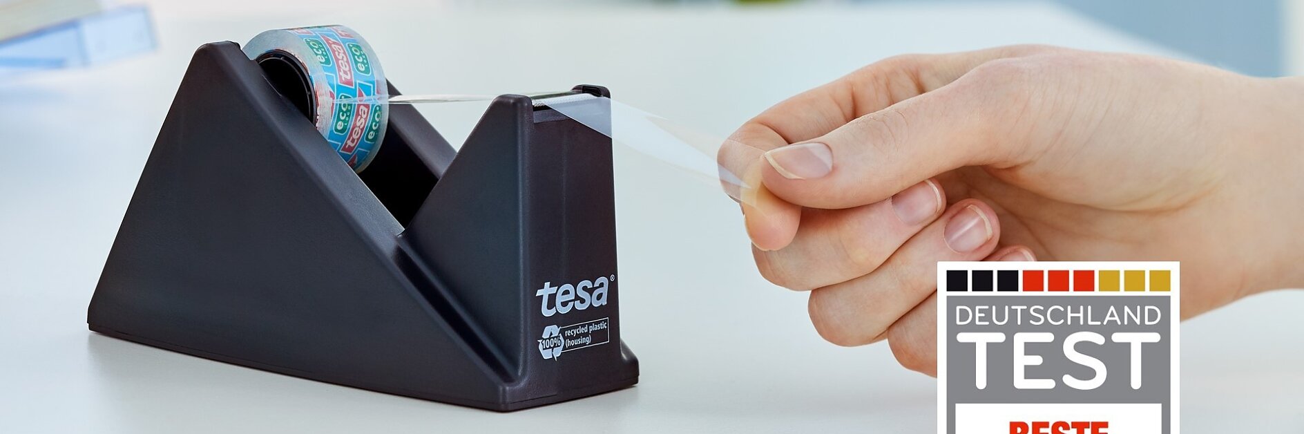 Eine Hand zieht einen Streifen Tesa-Klebeband aus einem schwarzen Klebebandspender mit der Aufschrift „Tesa“. Im Vordergrund ist eine Grafik des Deutschland Test-Preises mit der Auszeichnung „Beste Markenqualität“ zu sehen. Ein unscharfer weißer Hintergrund lässt eine Inneneinrichtung vermuten. (This text has been generated by AI)