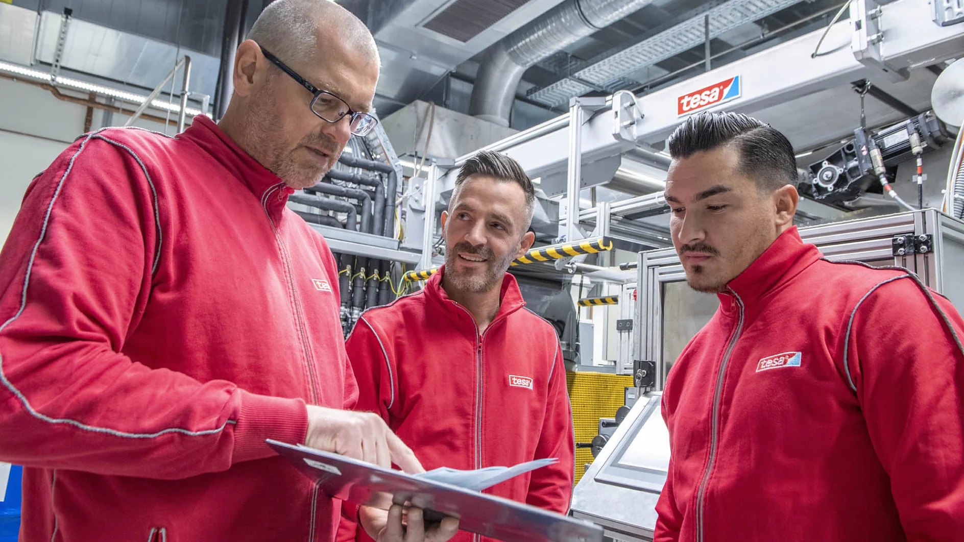 Drei Männer in roten Jacken stehen in einer Fabrik. Einer von ihnen bespricht etwas mit den anderen beiden und hält ein Klemmbrett in der Hand. Sie sind umgeben von Industriemaschinen und sichtbaren Rohrleitungen an der Decke, die alle sorgfältig mit Tesa-Klebeband angebracht wurden. (This text has been generated by AI)