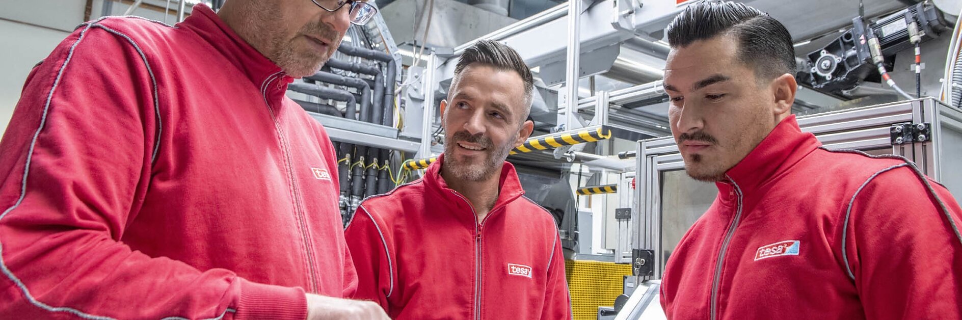 Drei Männer in roten Jacken stehen in einer Fabrik. Einer von ihnen bespricht etwas mit den anderen beiden und hält ein Klemmbrett in der Hand. Sie sind umgeben von Industriemaschinen und sichtbaren Rohrleitungen an der Decke, die alle sorgfältig mit Tesa-Klebeband angebracht wurden. (This text has been generated by AI)