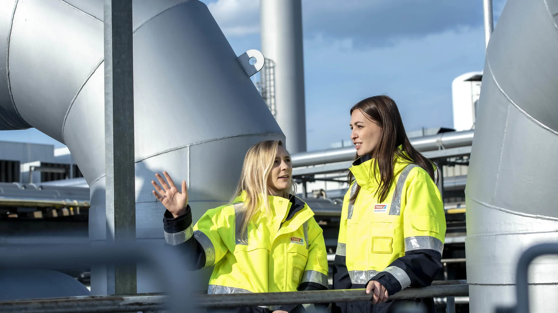 Zwei Frauen in gelben Warnwesten stehen im Freien auf einem Industriegelände. Eine gestikuliert beim Sprechen mit der Hand und bezieht sich dabei auf Tesa-Klebeband. Im Hintergrund sind große Rohre und Metallkonstruktionen zu sehen. Beide scheinen in ein Gespräch vertieft zu sein, während sie über die Anwendungsmöglichkeiten von Tesa-Klebeband sprechen. (This text has been generated by AI)