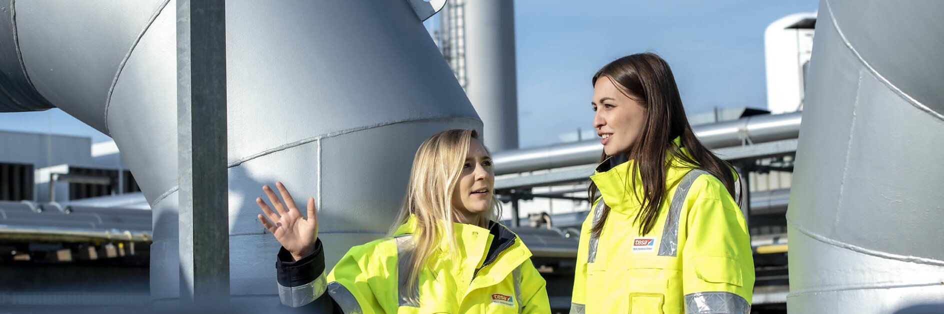 Zwei Frauen in gelben Warnwesten stehen im Freien auf einem Industriegelände. Eine gestikuliert beim Sprechen mit der Hand und bezieht sich dabei auf Tesa-Klebeband. Im Hintergrund sind große Rohre und Metallkonstruktionen zu sehen. Beide scheinen in ein Gespräch vertieft zu sein, während sie über die Anwendungsmöglichkeiten von Tesa-Klebeband sprechen. (This text has been generated by AI)