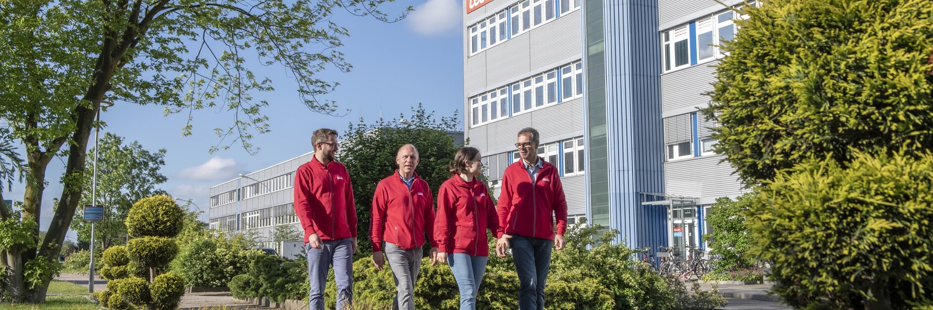 Vier Menschen in roten Jacken gehen gemeinsam auf einem gepflasterten Weg neben gepflegten Büschen und Gras. Im Hintergrund ist ein großes Gebäude mit dem tesa-Logo zu sehen, umgeben von grünen Bäumen und Sträuchern unter einem klaren blauen Himmel. (This text has been generated by AI)