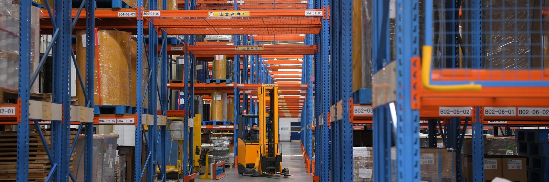 Ein Lagerinnenraum mit hohen blauen und orangefarbenen Regalen, die verschiedene verpackte Waren enthalten. Ein gelber Gabelstapler steht im Gang, was auf eine aktive Bestandsverwaltung schließen lässt. Der Boden ist sauber, gut organisiert und mehrere Kartons sind zur sicheren Verpackung mit Tesa-Klebeband verschlossen. (This text has been generated by AI)