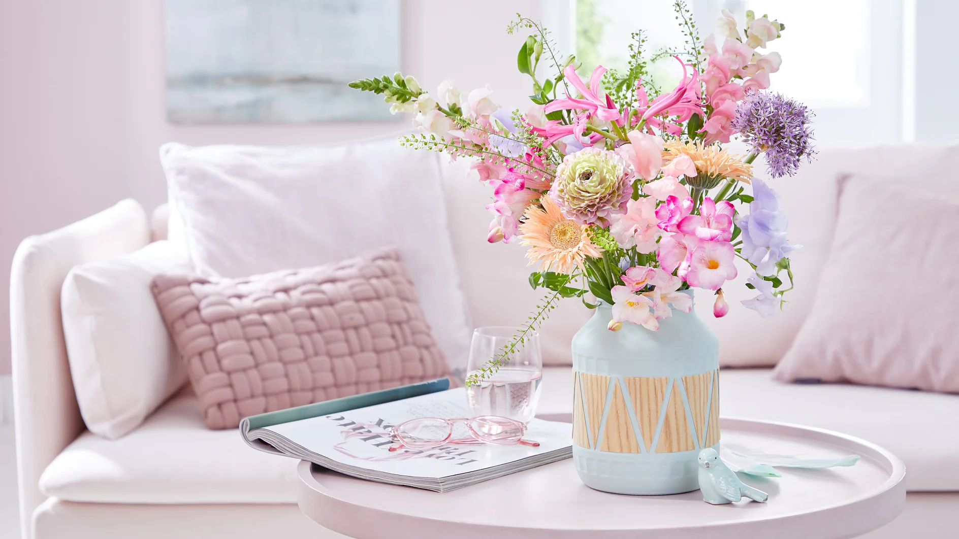 A pastel-colored living room with a round table featuring a vase of assorted flowers, an open magazine, a pair of glasses, and a decorative bird sculpture. A white sofa adorned with pink cushions is in the background. (This text has been generated by AI)