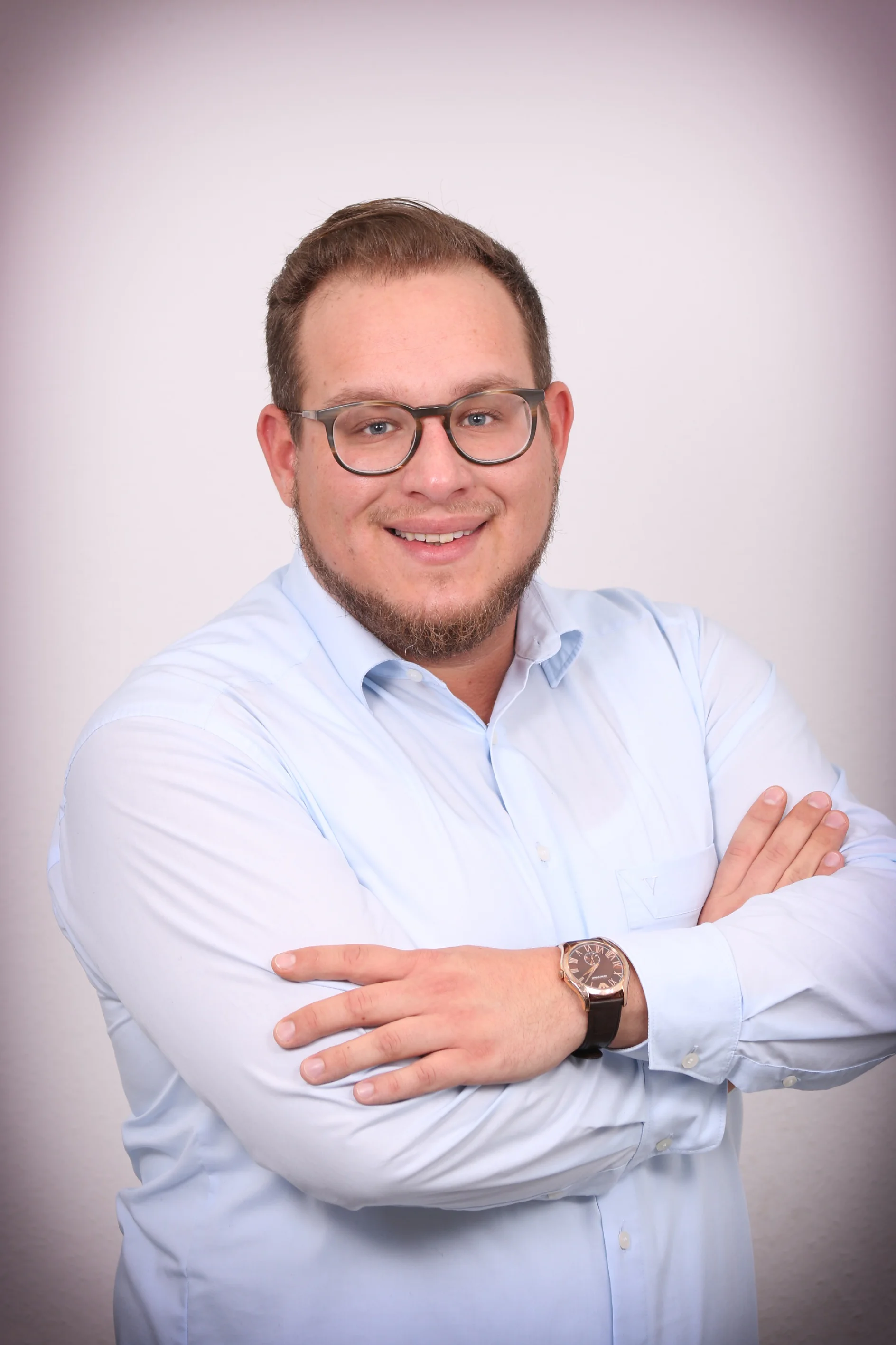 Ein Mann mit kurzen Haaren und Bart lächelt mit verschränkten Armen. Er trägt eine Brille, ein hellblaues Hemd mit Knöpfen und eine Uhr am linken Handgelenk. Der Hintergrund ist eine schlichte, cremefarbene Wand, die mit Rollen Tesa-Klebeband dekoriert ist. (This text has been generated by AI)
