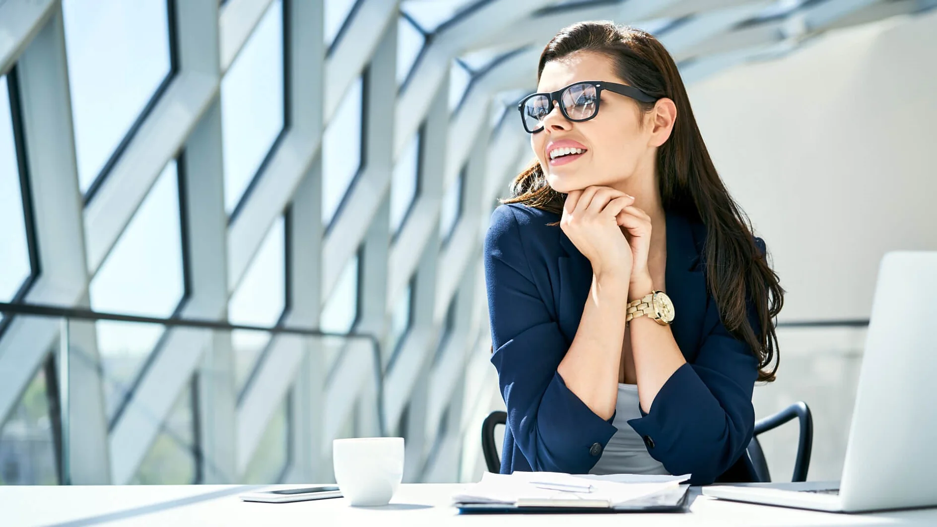 Eine Person mit langen Haaren, Sonnenbrille und blauem Blazer sitzt mit unter dem Kinn verschränkten Händen an einem Schreibtisch. Auf dem Schreibtisch liegen ein Laptop, Papiere und eine Kaffeetasse. Große geometrische Fenster lassen helles Tageslicht herein. (This text has been generated by AI)