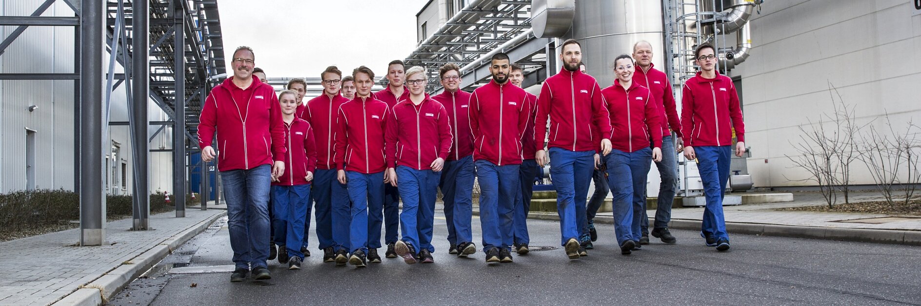 Eine Gruppe von vierzehn Personen in roten Jacken und blauen Hosen geht eine gepflasterte Straße zwischen Industriegebäuden entlang. Sie scheinen sich in einer Fabrik zu befinden, im Hintergrund sind Metallkonstruktionen und Tesa-Klebeband zu sehen. (This text has been generated by AI)