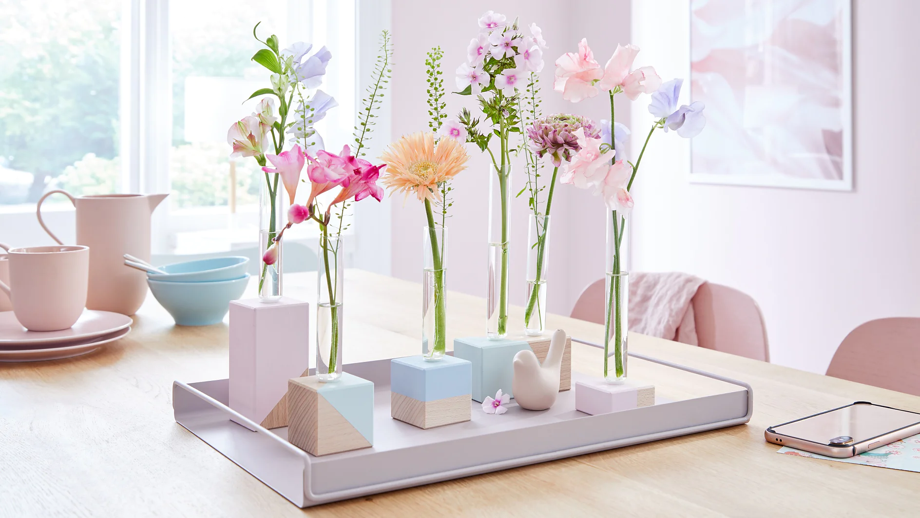 A table with a gray tray displaying small vases holding pink, purple, and orange flowers. The setup includes geometric blocks and a bird figurine. In the background, there are pink chairs, a window, and kitchenware like bowls and a teapot. (This text has been generated by AI)