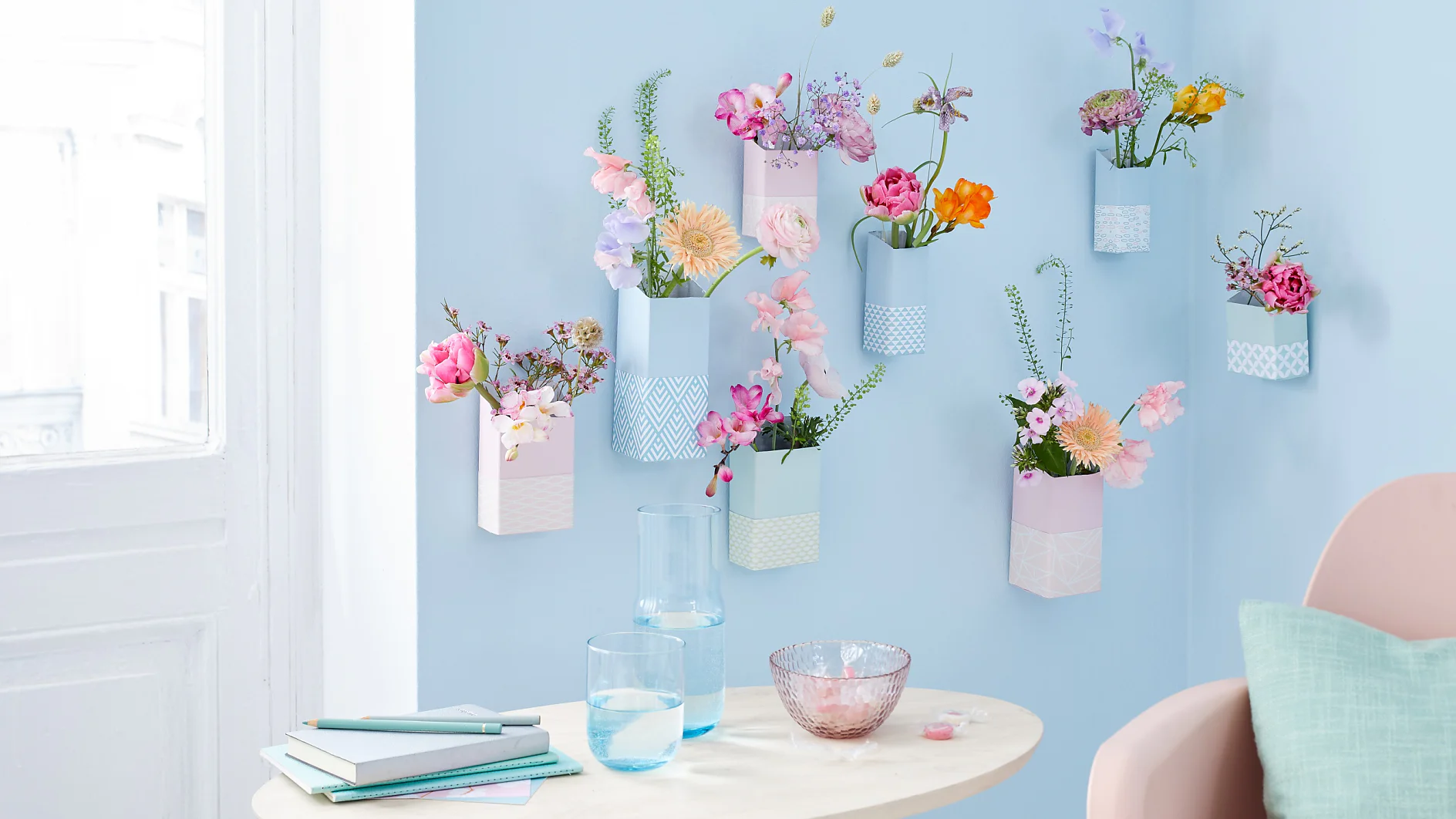 A light blue wall with geometric vases holding various colorful flowers, creating a vertical garden. A round wooden table below holds books, a notebook, two glasses of water, and a pink glass bowl. A pastel chair is in the foreground. (This text has been generated by AI)
