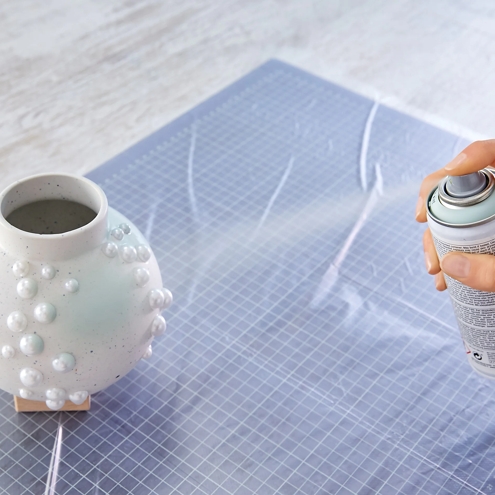 A hand holds a can of spray paint near a decorative vase with raised circular patterns. The vase rests on a protective mat on a table, secured with tesa tape. (This text has been generated by AI)