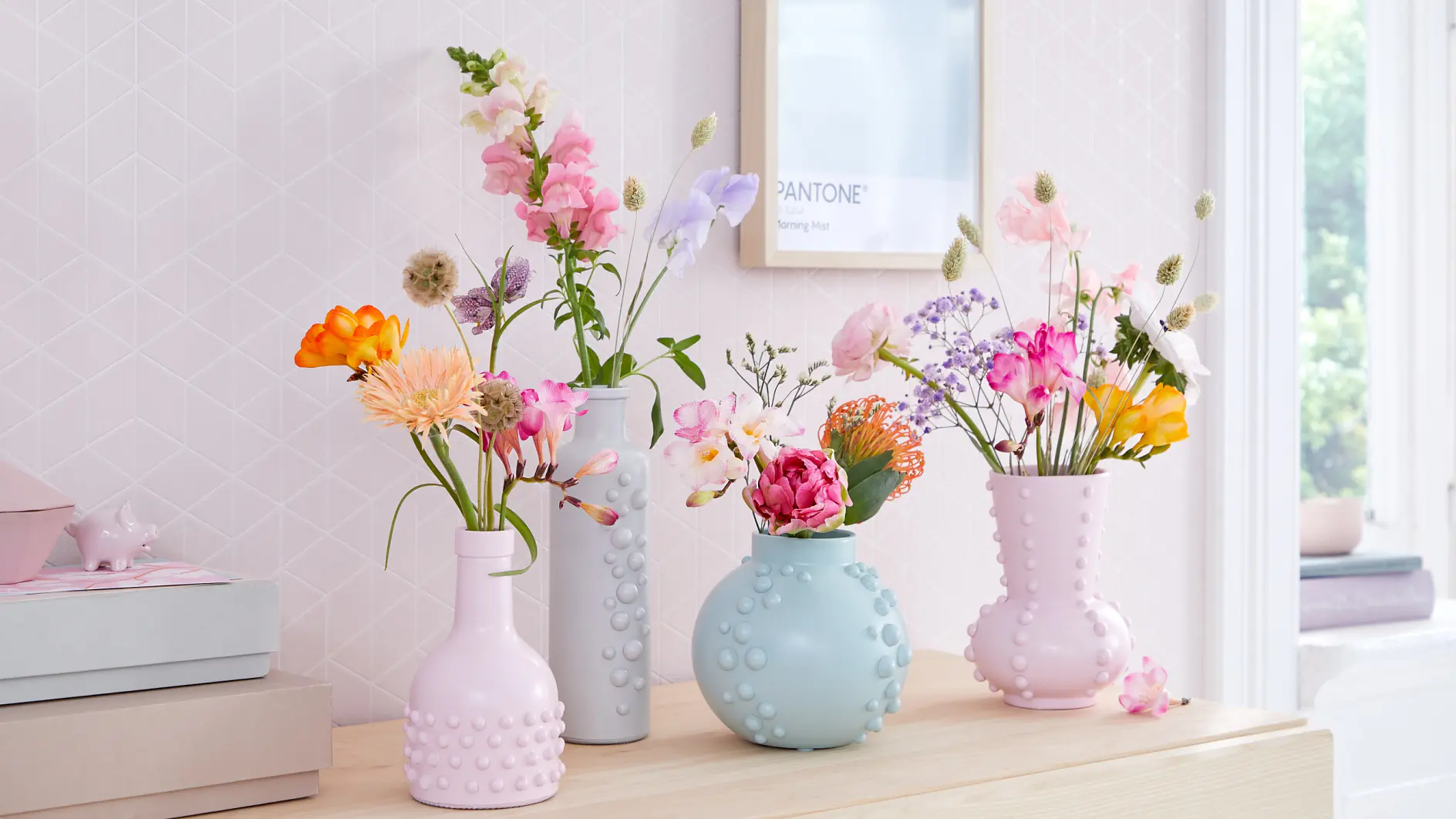 Four pastel-colored vases with textured designs sit on a wooden surface, each holding a variety of colorful flowers. The background features a light-colored wall with a framed picture and soft natural light from a nearby window.